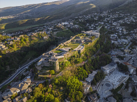 Aerial view of Gjirokaster Fortress perched on a hill overlooking the historic stone city with lush green trees and surrounding mountains in Gjirokaster, Gjirokaster County, Albania.