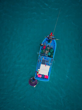 Aerial view of a small blue fishing boat with a round basket boat attached, carrying fishing nets and a solar panel on the deck, floating in turquoise sea water in Vietnam.