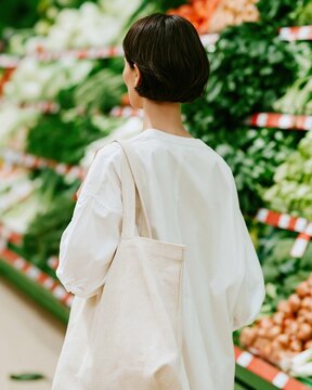 Woman from behind carrying a reusable cloth tote bag while shopping for fresh vegetables in a grocery store for sustainable lifestyle and mindful living themes