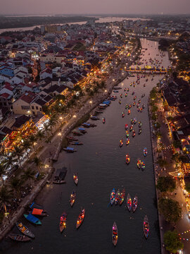 Aerial view of the Thu Bon River in Hoi An at dusk, featuring numerous traditional wooden boats with glowing lanterns, historic yellow buildings, and palm trees lining the riverbanks in Vietnam.