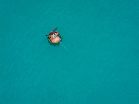 Aerial view of a traditional round basket boat with a fisherman floating on the clear turquoise water of the sea in Vietnam.