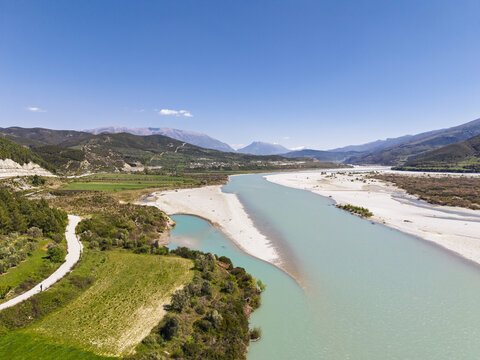 Aerial view of Vjosa Wild River National Park with its turquoise river, gravel banks, and surrounding green hills under a clear blue sky in Permet, Gjirokaster County, Albania.