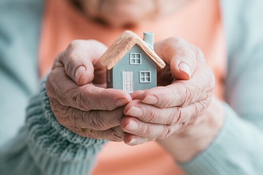 Wrinkled elderly hands cradling a miniature house model with a textured roof, showing weathered skin and soft knit textures for home insurance and mortgage concepts