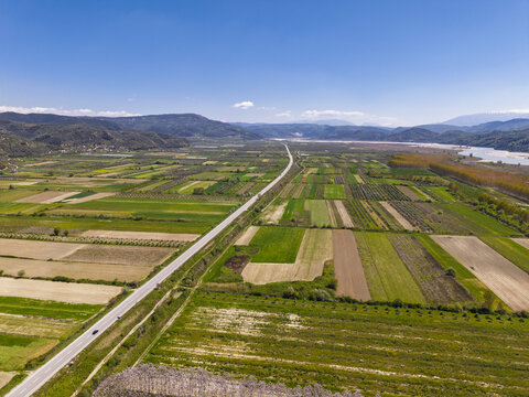 Aerial view of agricultural fields, a long straight road cutting through the landscape, and distant mountains under a clear blue sky in Lushnje, Fier County, Albania.