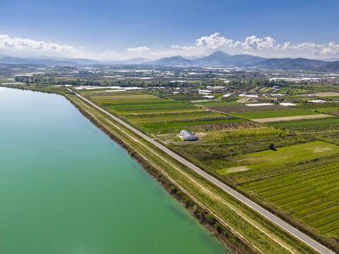 Aerial view of agricultural fields and Liqeni Thanes turquoise reservoir with distant mountains under a clear blue sky in Lushnje, Fier County, Albania.