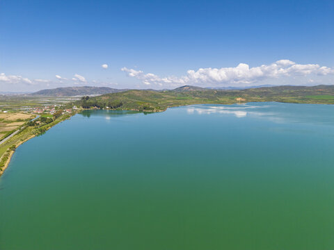 Aerial view of Liqeni Thanes turquoise lake surrounded by rolling hills, green fields, and a clear blue sky in Lushnje, Fier County, Albania.
