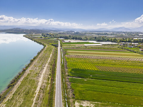 Aerial view of a road running alongside Liqeni Thanes lake and vibrant agricultural fields with greenhouses under a clear blue sky, Lushnje, Fier County, Albania.
