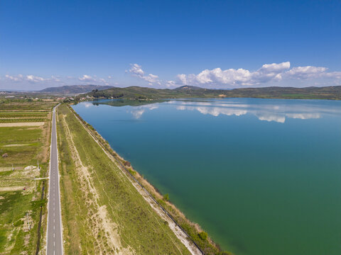 Aerial view of Liqeni Thanes serene reservoir with turquoise water reflecting the blue sky, bordered by a long asphalt road and green agricultural fields in Lushnje, Fier County, Albania.
