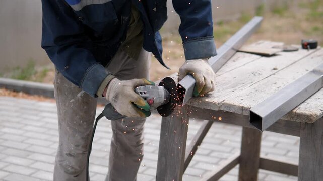 Worker in dusty workwear and gloves cuts square metal tubing with handheld angle grinder beside weathered outdoor workbench, sending bright sparks downward through muted daylight and gritty