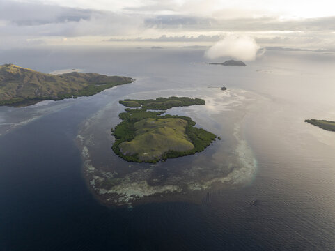 Aerial view of Komodo Island surrounded by turquoise coral reefs and deep blue waters under a soft cloudy sky in Komodo, East Nusa Tenggara, Indonesia.