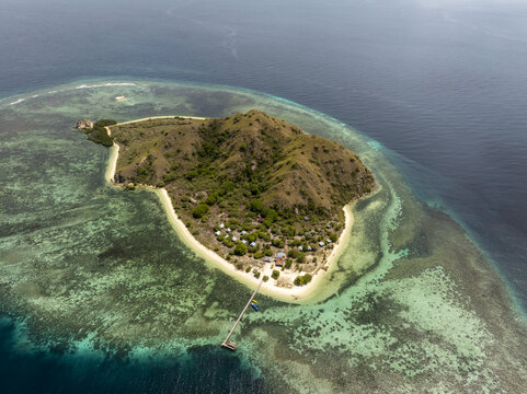 Aerial view of Kanawa Island with lush green hills, white sandy beaches, and a wooden pier extending into turquoise coral reef waters in Kecamatan Komodo, Nusa Tenggara Timur, Indonesia.