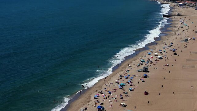 Aerial view of Will Rogers State Beach with people relaxing under umbrellas on the sand and waves breaking along the shoreline in Los Angeles, California, United States.