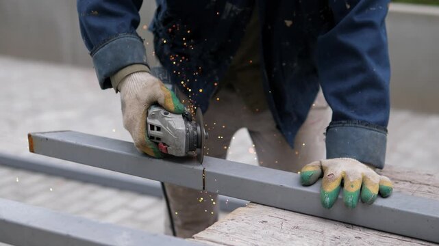 Gloved worker in blue workwear uses handheld grinder on metal beam as bright sparks fly from cutting point in rugged workshop setting, captured from tight waist-level view with blurred background