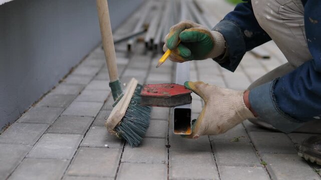 Crouched worker in protective gloves holds yellow pencil against metal tube beside brush and broom on gray pavers, suggesting careful measuring or fitting during outdoor repair work with soft