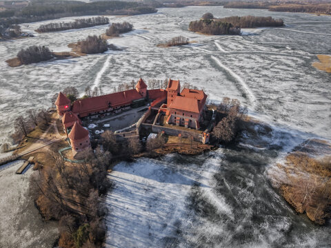 Aerial view of Trakai Island Castle, a historic red-brick fortress on an island in Lake Galve, surrounded by frozen water and bare trees in Trakai, Vilnius County, Lithuania.