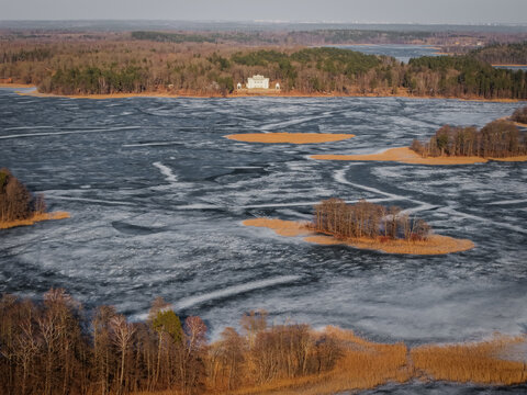 Aerial view of the historic Uzutrakis Manor situated on the shore of a frozen lake surrounded by dense forest and ice patterns in Trakai, Vilnius County, Lithuania.