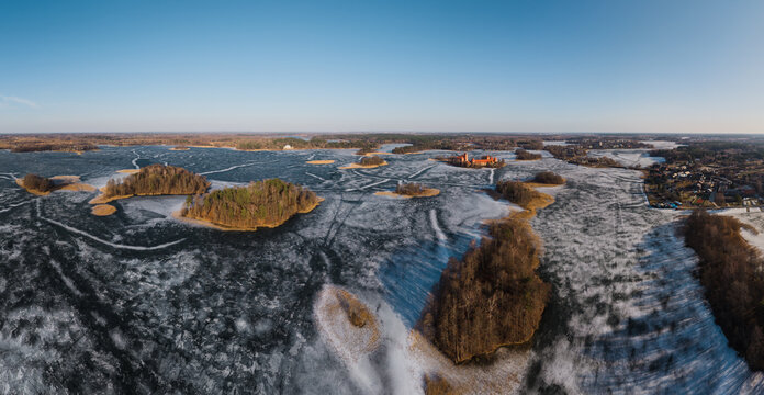 Aerial view of Trakai Island Castle situated on a frozen lake with ice patterns, surrounded by small wooded islands under a clear blue sky in Trakai, Vilnius County, Lithuania.