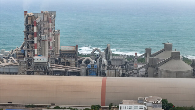 Aerial view of a large industrial cement factory complex with tall silos, complex piping systems, and conveyor belts situated along the coastline in Indonesia.