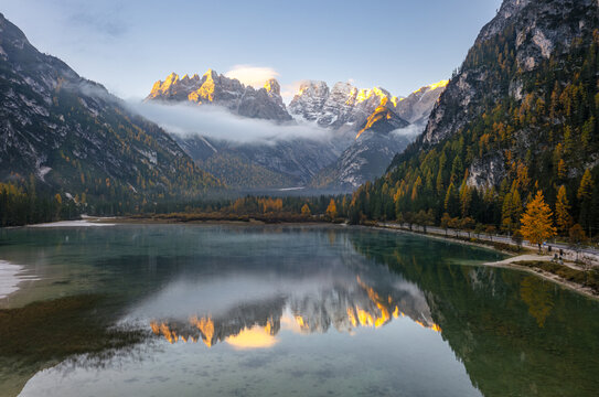 Aerial view of Durrensee reflecting the jagged, snow-dusted peaks of the Dolomites surrounded by vibrant autumn foliage and clear turquoise water in Toblach, Trentino-Alto Adige, Italy.
