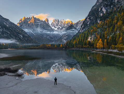 Aerial view of a person standing on the shore of Durrensee lake reflecting snow-capped Dolomites peaks during golden hour with autumn foliage in Toblach, Trentino-Alto AdigeS, Italy.