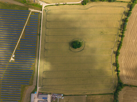 Aerial view of a solar farm with rows of photovoltaic panels adjacent to a large agricultural field featuring a central cluster of trees in Melton Mowbray, England, United Kingdom.