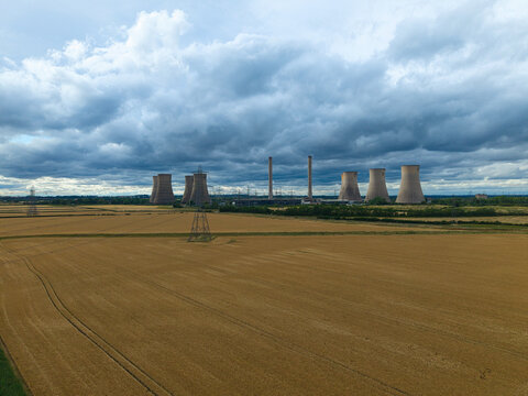 Aerial view of West Burton Power Station with its iconic cooling towers rising above golden wheat fields under a dramatic cloudy sky in West Burton, England, United Kingdom.