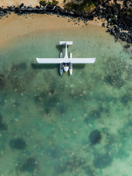 Aerial view of a white seaplane floating in the clear turquoise shallow water near a sandy beach and rocky coastline in Mauritius.