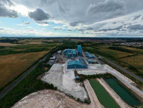 Aerial view of the Saint Gobain Formula industrial plant, featuring blue-roofed buildings, gypsum stockpiles, and sediment ponds surrounded by rural fields in New Balderton, England, United Kingdom.