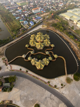 Aerial view of the Pavilion of the Enlightened, a golden structure set in a reflective pond with intricate walkways, surrounded by urban landscape in Phraeksa, Samut Prakan, Thailand.
