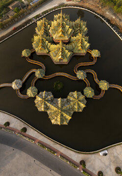 Aerial view of the Pavilion of the Enlightened, featuring golden tiered roofs surrounded by a reflective pond and connected by winding walkways in Phraeksa, Samut Prakan, Thailand.
