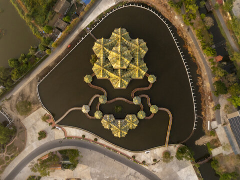 Aerial view of the Pavilion of the Enlightened, a golden structure surrounded by water, connected by wooden walkways, set within a park in Phraeksa, Samut Prakan, Thailand.