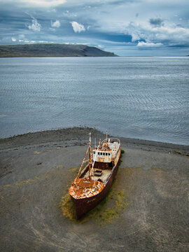 Aerial view of the rusted shipwreck Gardar BA 64 resting on a gravel beach by the sea under a cloudy sky in Saudlauksdalur, Vesturbyggd, Iceland.