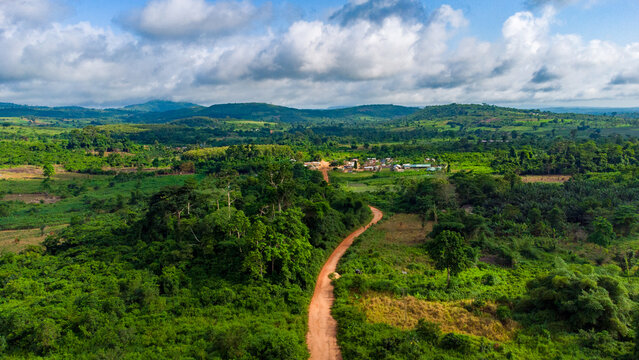 Aerial view of the lush green landscape and rolling hills of Aburi with a winding dirt road leading to a small village under a cloudy sky in Aburi, Eastern Region, Ghana.