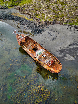 Aerial view of the rusted Stykkisholmur Shipwreck resting in shallow clear water near a rocky shoreline in Breidhabolsstadhur, Snaefellsnes, Iceland.