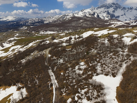 Aerial view of Durmitor National Park with snow-capped mountains, winding dirt roads, patches of snow on the landscape, and sparse forest in Zabljak, Zabljak Municipality, Montenegro.