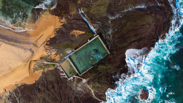 Aerial view of the Mona Vale Rock Pool situated on a rocky coastline with turquoise ocean waves crashing against the shore and golden sand in Mona Vale, New South Wales, Australia.