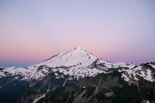 View of the snow-capped peak of Mount Baker at Artist Point with rugged slopes, evergreen forests, and a soft purple twilight sky in Deming, Washington, United States.