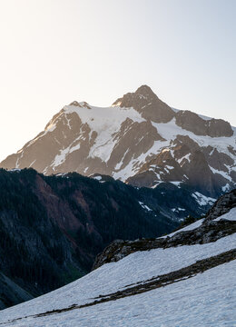 View of the snow-capped peak of Mount Shuksan illuminated by soft golden hour light, with rugged mountain slopes and patches of snow in the foreground in Deming, Washington, United States.