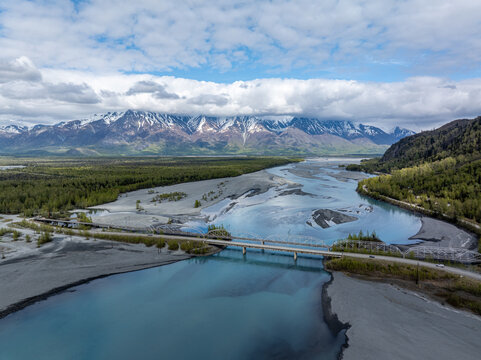 Aerial view of the Knik River bridge spanning the glacial river channels with snow-capped mountains in the background under a cloudy sky in Knik River, Alaska, United States.