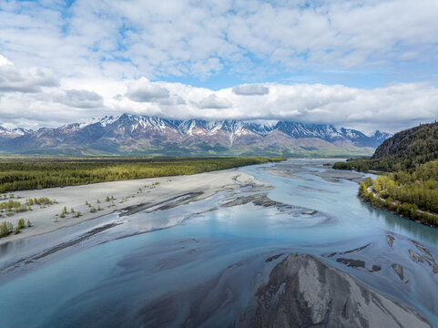 Aerial view of Knik River with snow-capped mountains, lush forests, and gravel riverbeds under a cloudy sky in Knik River, Alaska, United States.