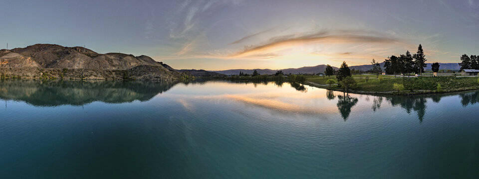 Aerial view of the calm waters of Lake Dunstan reflecting the golden sunset light, surrounded by rolling hills and trees in Cromwell, Otago Region, New Zealand.