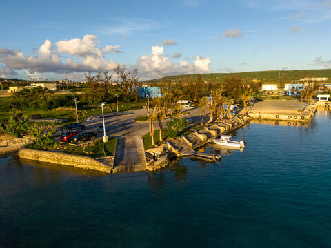 Aerial view of Tinian Port and marina with boats docked at the pier, palm trees, parked vehicles, and clear blue water in San Jose, Tinian, Northern Mariana Islands.