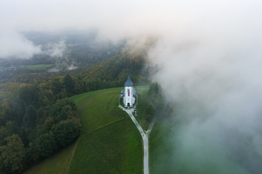 Aerial view of the Church of Saints Primus and Felician surrounded by green hills and autumn forests, partially obscured by thick morning mist in Jamnik, Kranj, Slovenia.