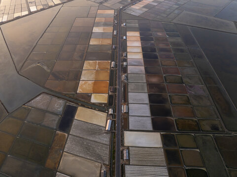 Aerial view of the geometric salt pans at Laem Phak Bia featuring varying shades of water and earth, separated by a central road, in Laem Phak Bia, Thailand.