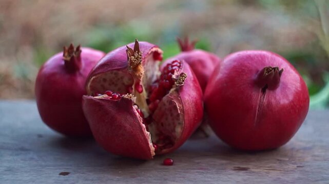 Slow motion camera panning of fresh red pomegranates with one cracked open revealing juicy seeds on a rustic wooden table, horizontal video.