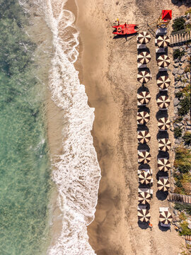 Aerial view of a sandy beach featuring rows of striped beach umbrellas, a red lifeguard boat, and turquoise water with gentle waves, Sardinia, Italy.