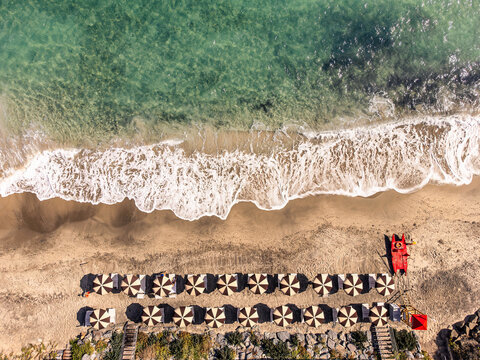 Aerial view of a sandy beach with rows of striped umbrellas, turquoise sea water, white waves, and a red lifeguard boat on the shore, Sardinia, Italy.