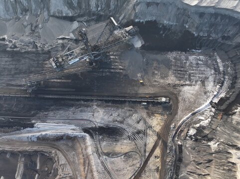 Aerial view of a massive bucket-wheel excavator operating in an open-pit coal mine with dark earth, conveyor belts, and industrial machinery in Germany.