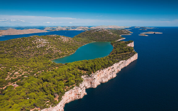 Aerial view of the salt lake Mir within Telascica Nature Park, surrounded by lush pine forests and dramatic limestone cliffs overlooking the Adriatic Sea in Sali, Zadar County, Croatia.