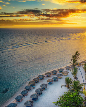 Aerial view of Cafe Del Sol beach with rows of thatched umbrellas on white sand along the calm sea during a vibrant golden hour sunset in Guayacanes, San Pedro de Macoris, Dominican Republic.